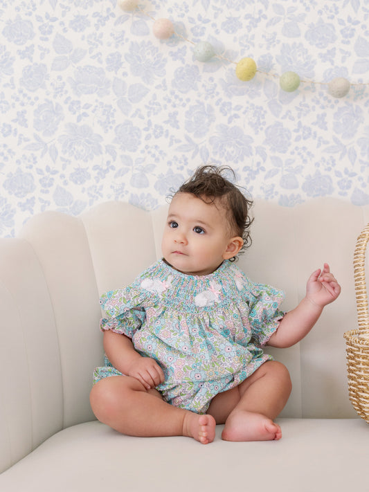 Baby sitting on a couch wearing a floral outfit with a light blue floral wallpaper background.