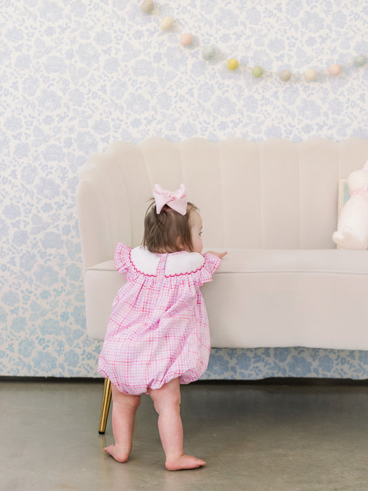 Child in a pink checkered dress standing in front of a beige sofa with decorative wall paper.