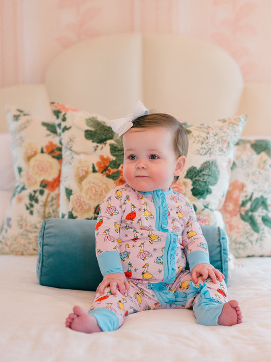 Baby in colorful romper with goose pattern sitting on a bed with floral pillows