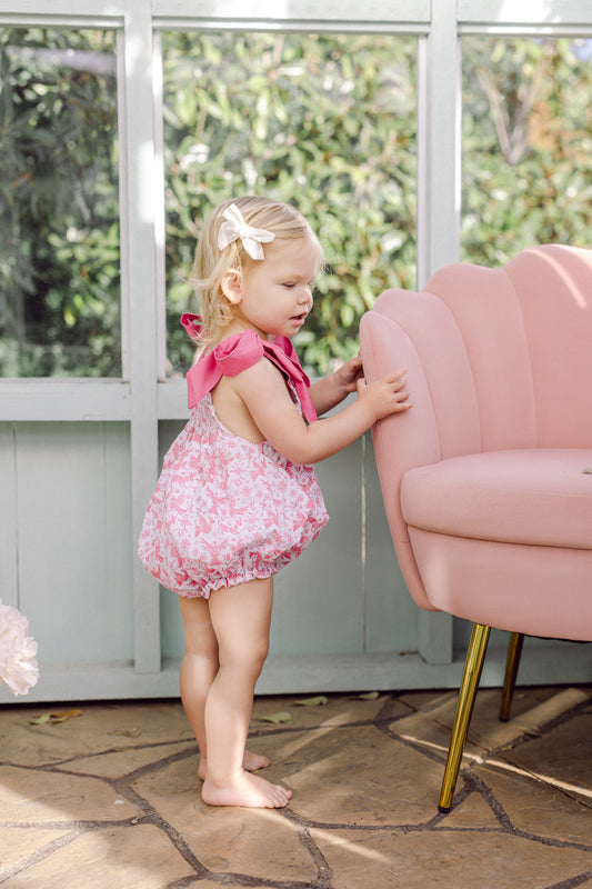 Toddler girl wearing a pink smocked floral bubble with bow straps, standing barefoot beside a blush chair indoors.