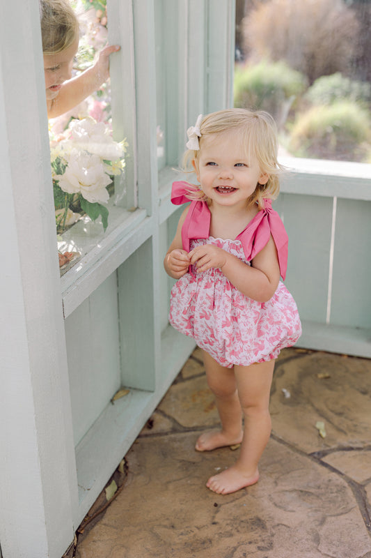 A young child wearing a pink smocked bubble with a mexican design and a bow on the sleeves standing near a window.