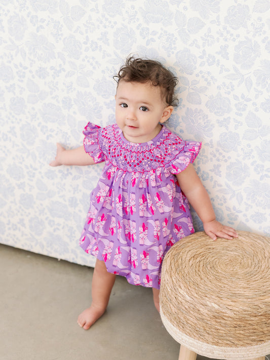 Baby in a purple dress with pink patterns standing against a textured white wall.