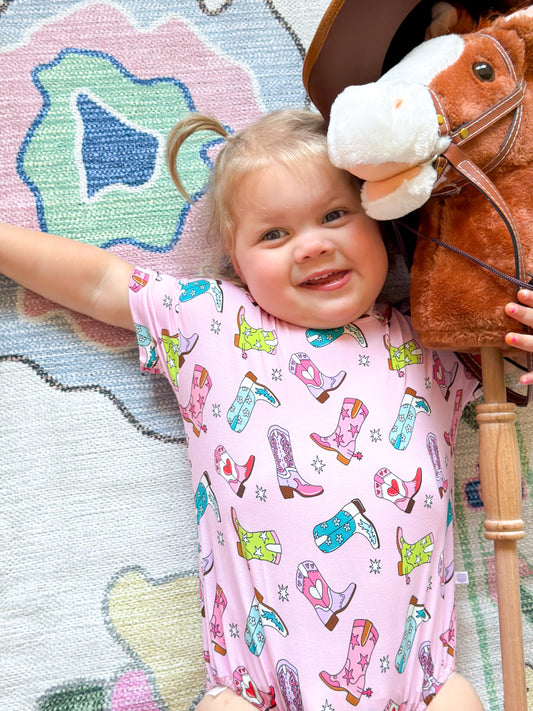 A toddler girl in a pink cowboy boot print bamboo t-shirt bubble holding a toy horse on a colorful rug.