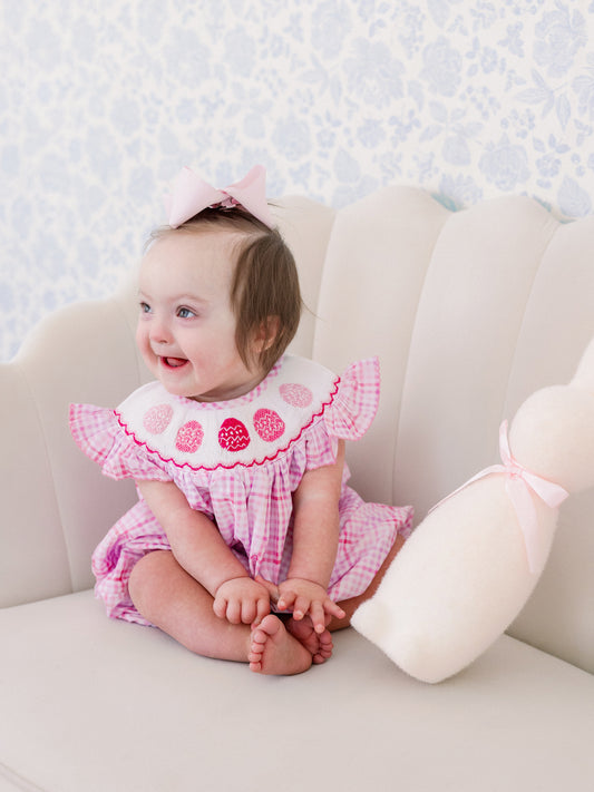 Baby in a pink easter egg outfit sitting on a white couch with a floral wallpaper background