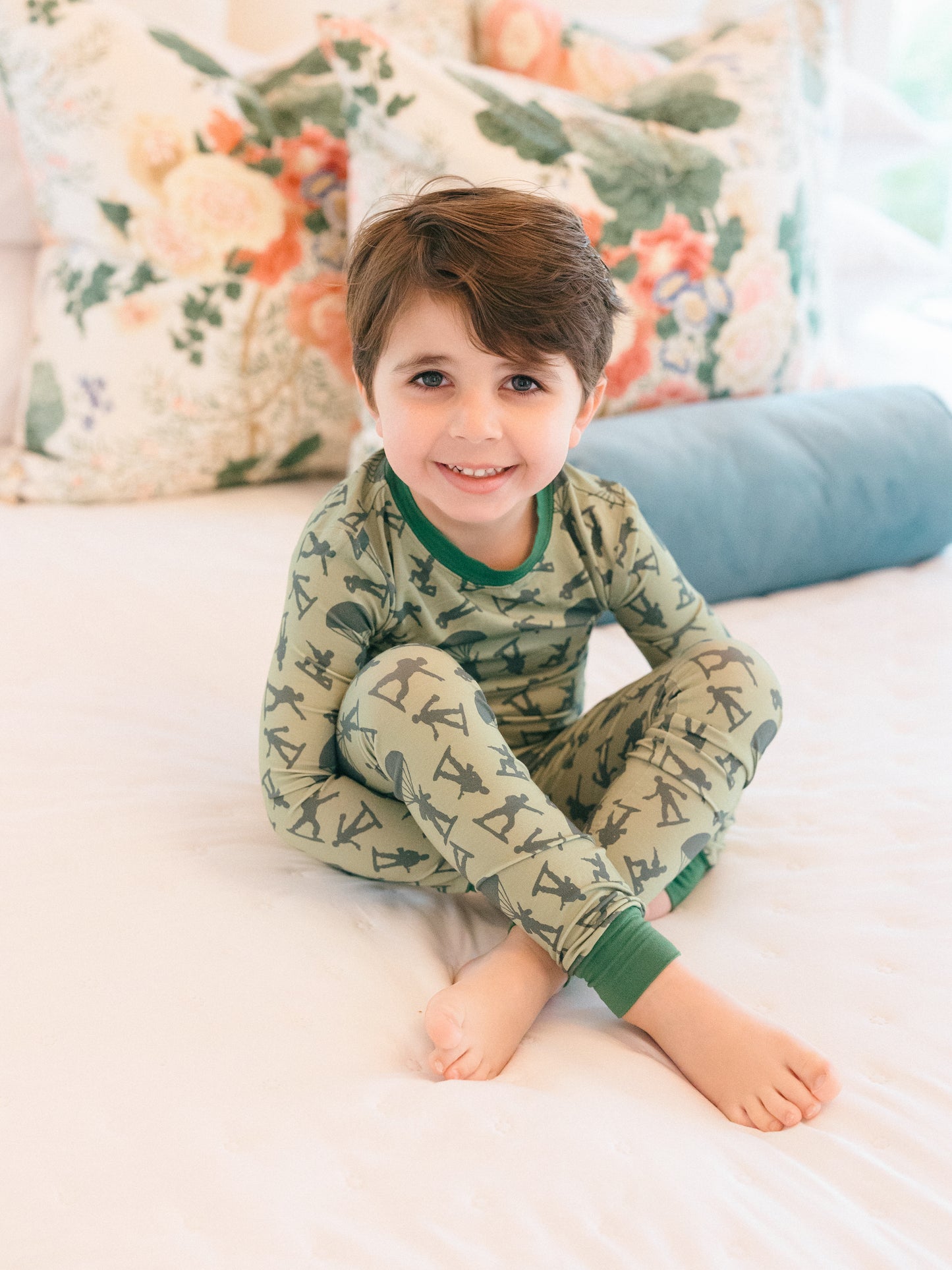 Child wearing green pajamas with an army man pattern, sitting on a bed with floral pillows.