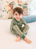 Child wearing green pajamas with an army man pattern, sitting on a bed with floral pillows.