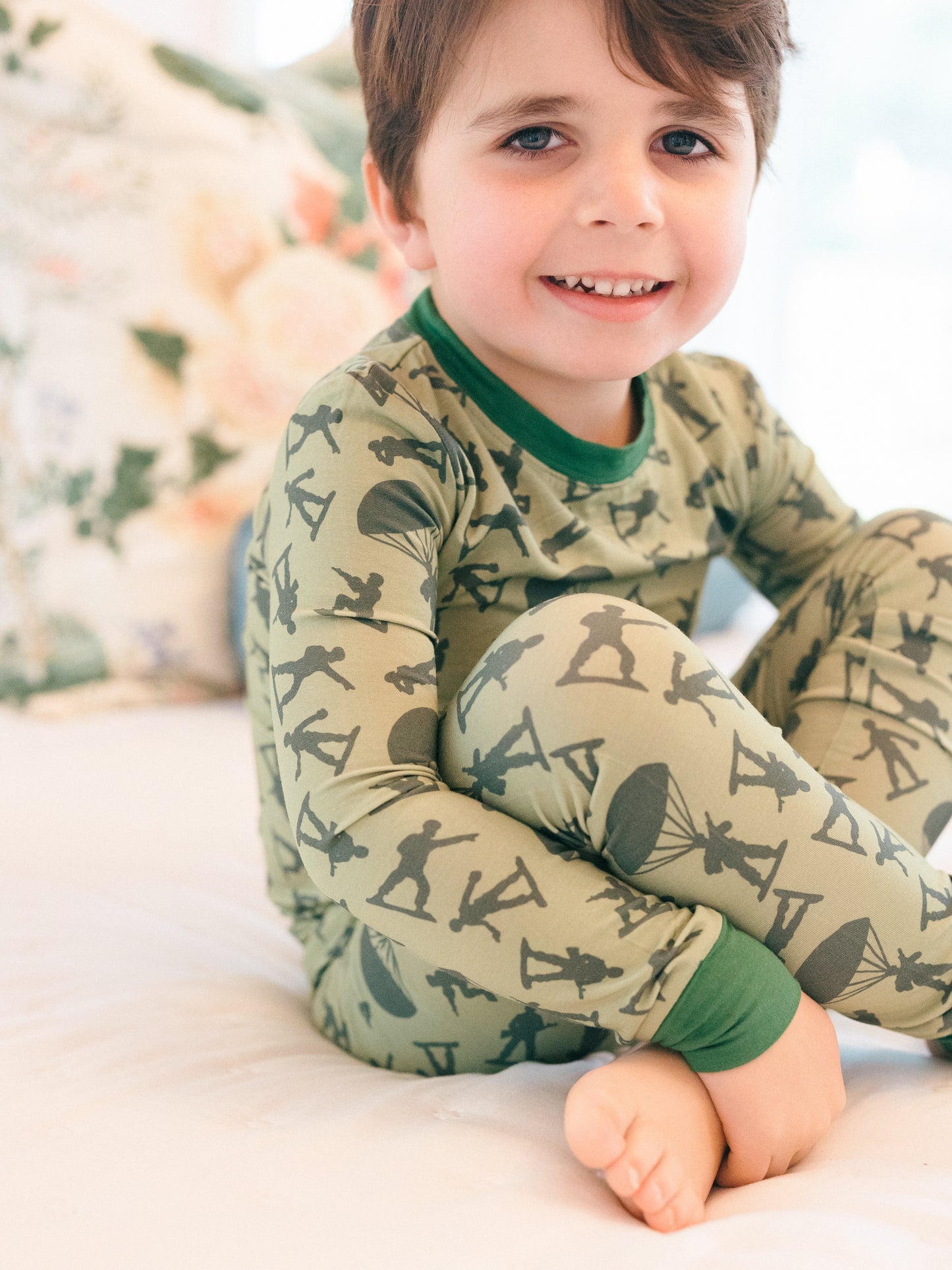 Child wearing a green army man patterned onesie sitting on a white surface.