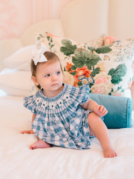 Baby in a blue checkered diaper cover set sitting on a bed with floral pillows in the background