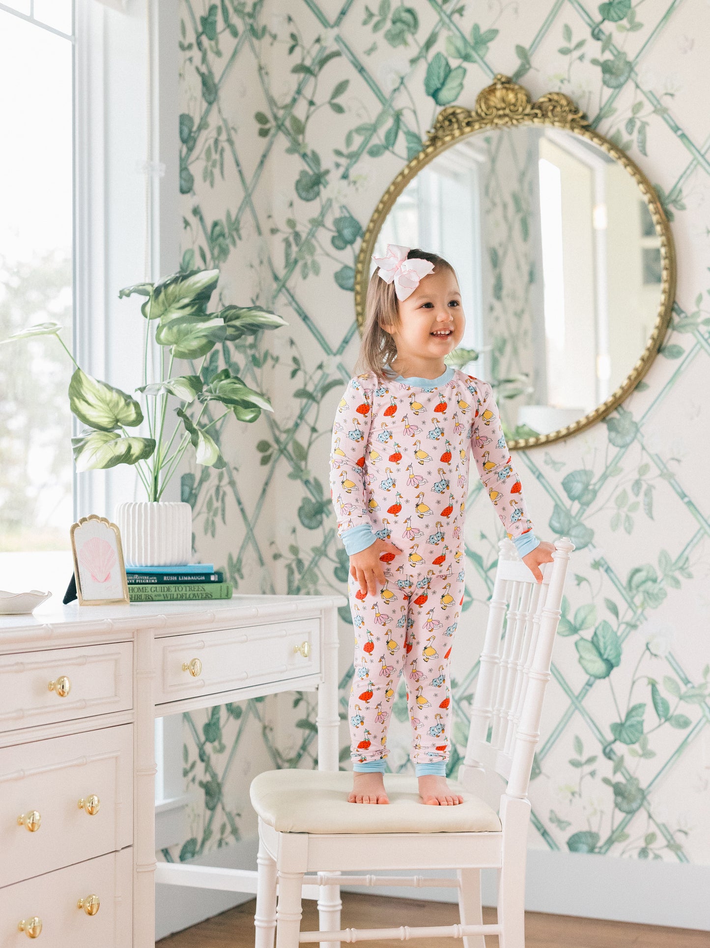 Child in a floral pj set with goose pattern standing on a chair in a room with decorative wallpaper and furniture.
