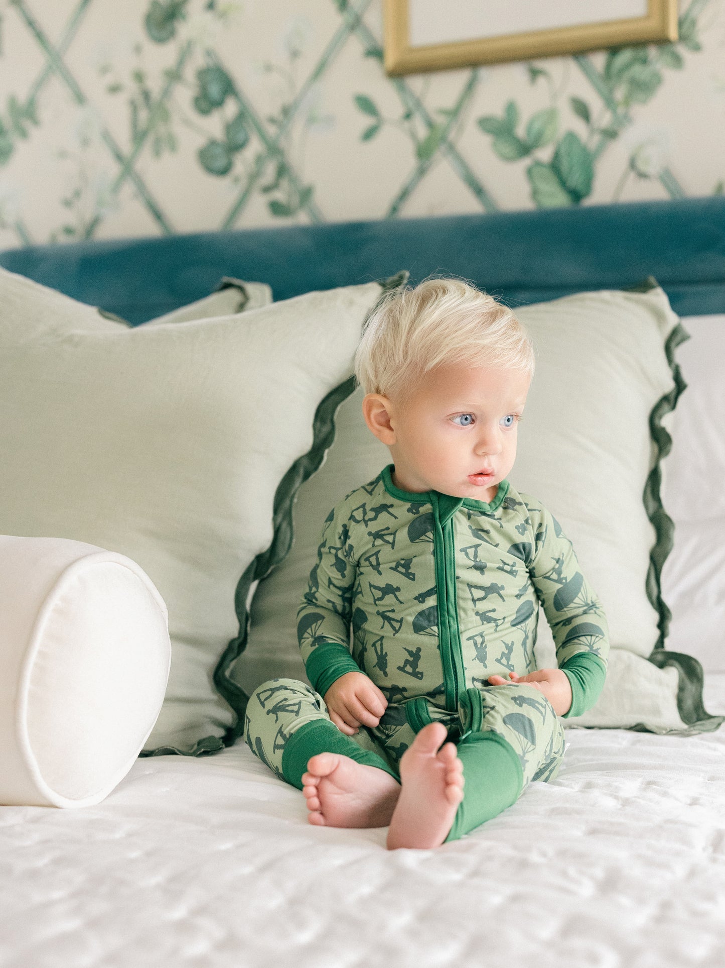 Baby in green pajamas sitting on a bed with floral wallpaper.