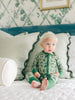 Child wearing green romoper with an army pattern, sitting on a bed with decorative pillows and wallpaper.
