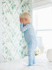 Child wearing a light blue duck onesie with a subtle pattern, standing in a room with white walls and a decorative curtain.