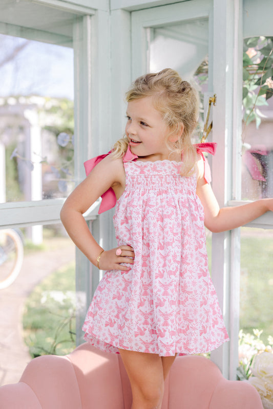 Little girl wearing a smocked pink and white bow tie dress standing by a window. 