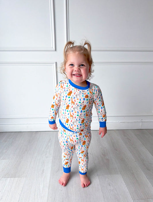 Child wearing colorful party animal pajamas standing on a light wooden floor against a white wall.