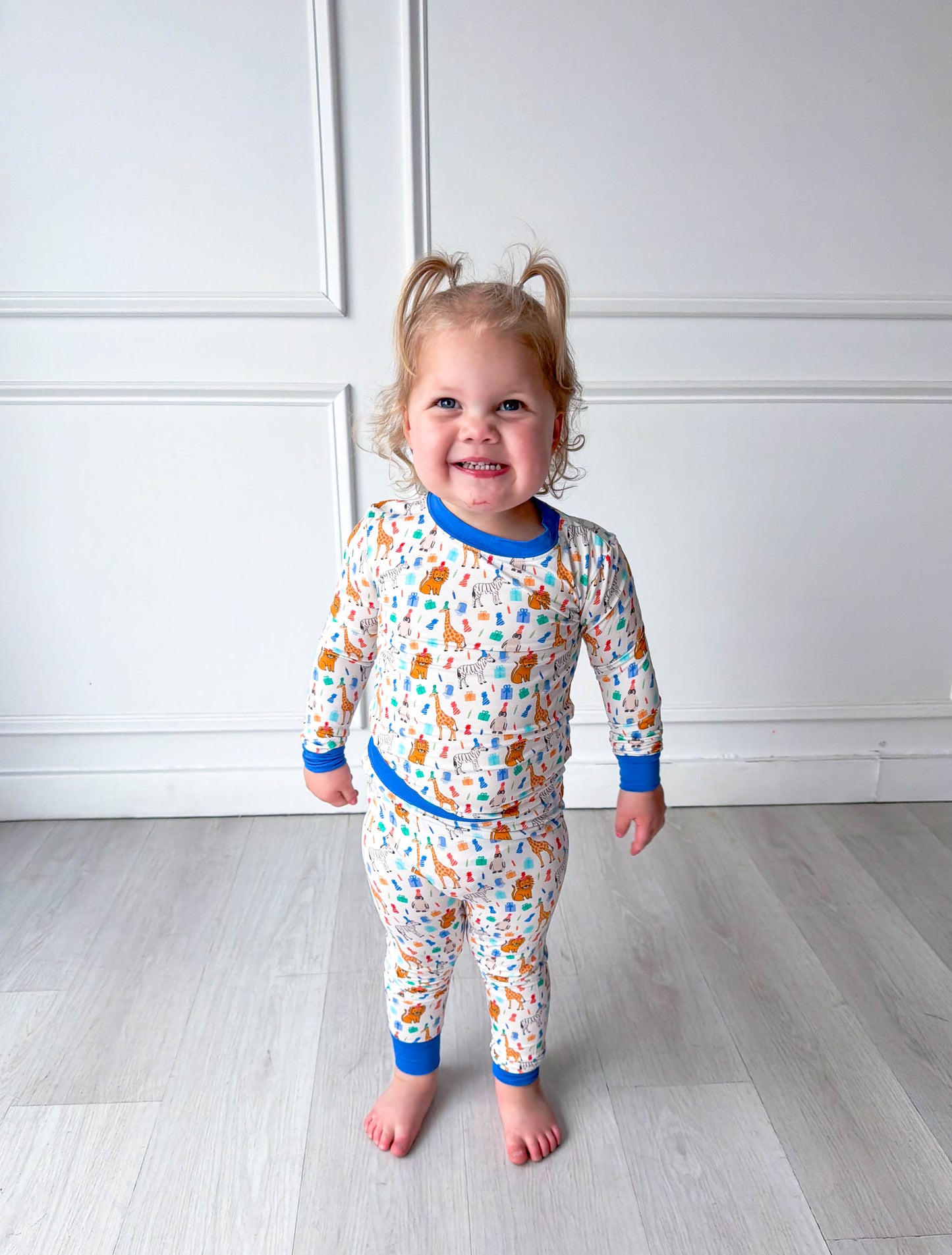 Child wearing colorful party animal pajamas standing on a light wooden floor against a white wall.