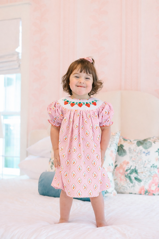 Young girl in a pink strawberry dress with floral patterns standing on a bed.