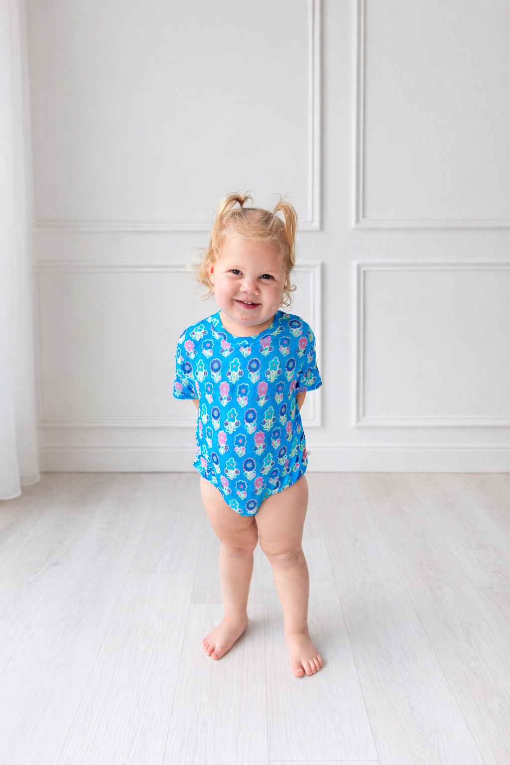 Toddler in blue floral t shirt bubble standing in front of a white wall. 