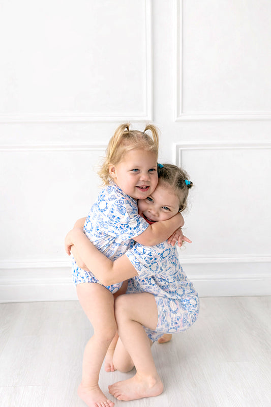 Two young girls in matching blue floral outfits hugging against a white wall.