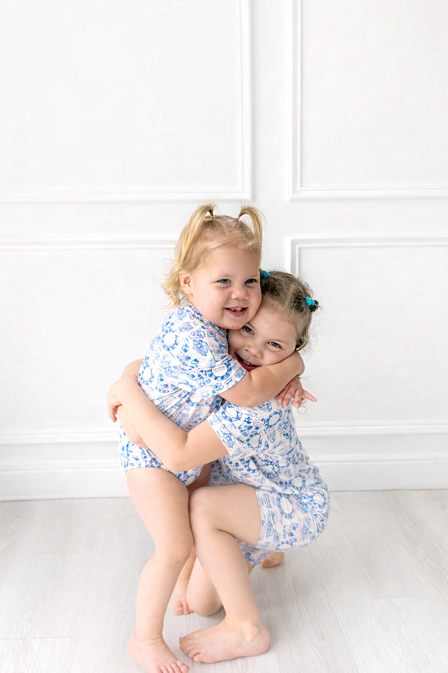 Two young girls in matching blue floral outfits hugging against a white wall.