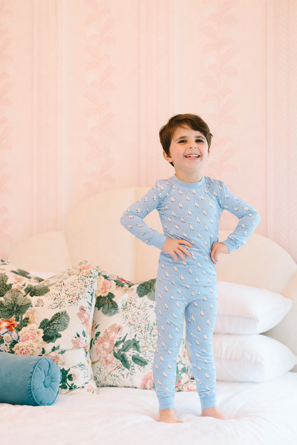 Child wearing a blue pajama set with a pattern of small geese, standing on a bed with floral pillows and white bedding.